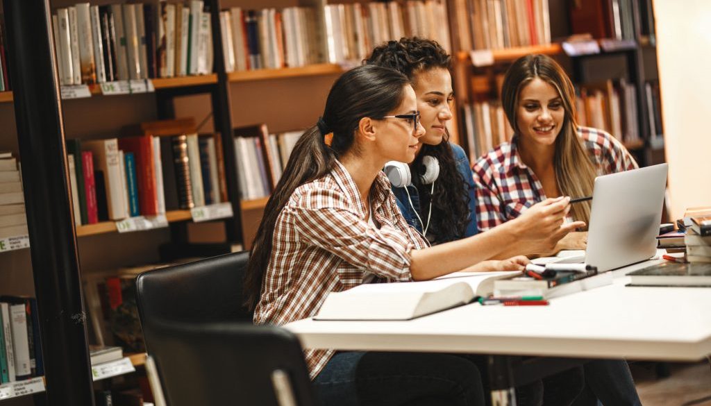 Three young women collaborate on a project in a library, utilizing a shared workspace. This highlights the convenience of reserving study areas with a space booking app for efficient teamwork and learning.