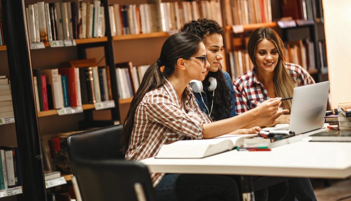 Three young women collaborate on a project in a library, utilizing a shared workspace. This highlights the convenience of reserving study areas with a space booking app for efficient teamwork and learning.