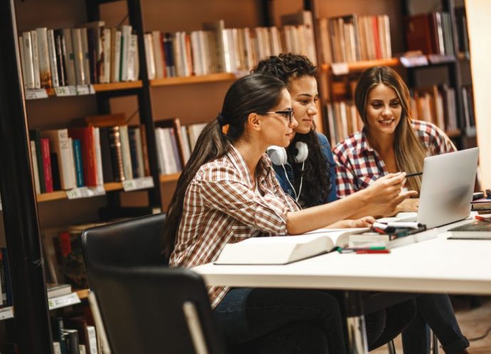 Three young women collaborate on a project in a library, utilizing a shared workspace. This highlights the convenience of reserving study areas with a space booking app for efficient teamwork and learning.