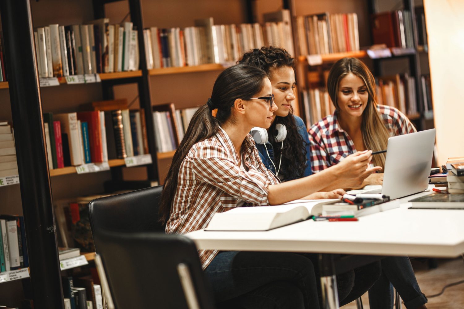 Three young women collaborate on a project in a library, utilizing a shared workspace. This highlights the convenience of reserving study areas with a space booking app for efficient teamwork and learning.