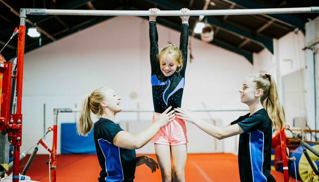 Two coaches assist a young gymnast practicing on the high bar in a brightly lit gym. The scene highlights the importance of safety, teamwork, and structured training programs, making it ideal for gymnastics management software that streamlines scheduling and class organization